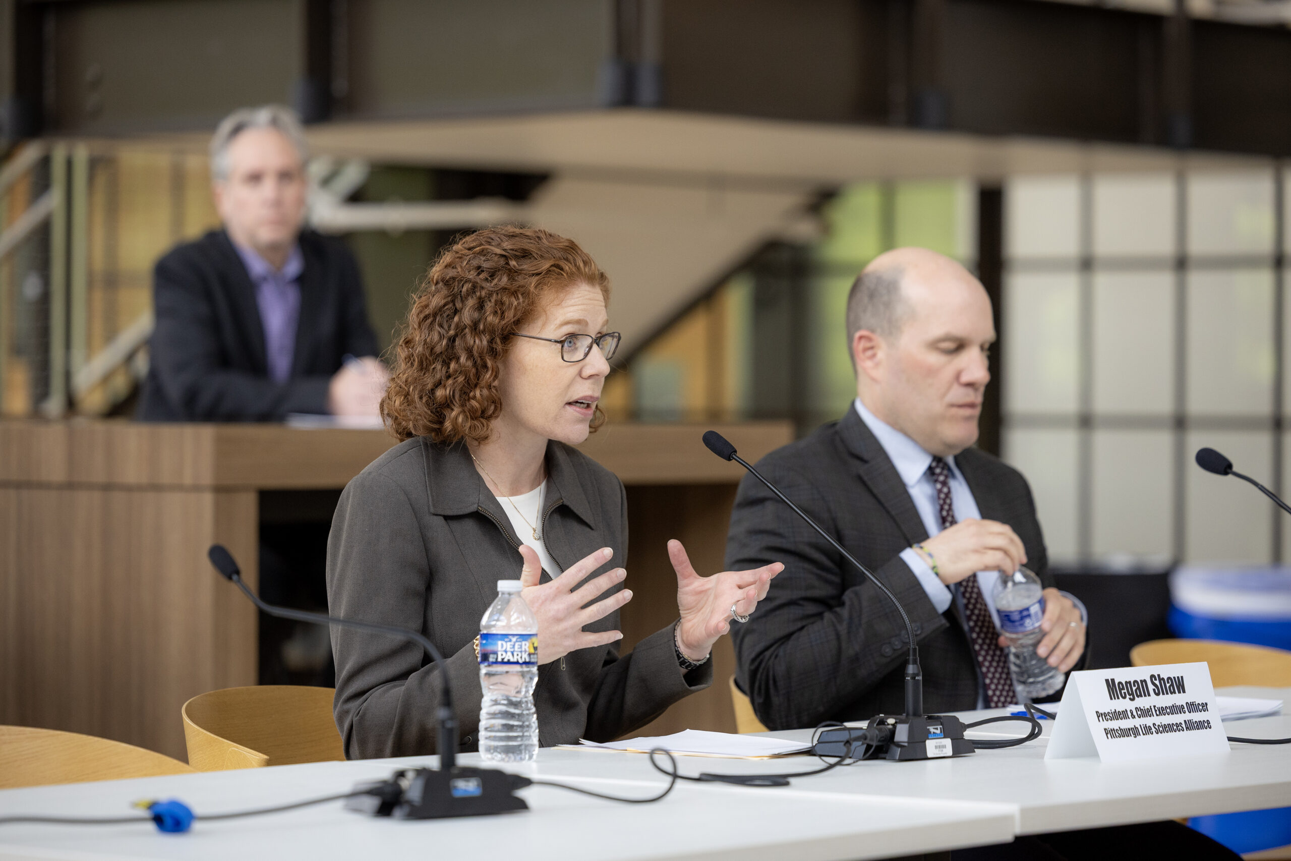 A woman and a man sit at a table with microphones and water bottles, speaking at a panel discussion; another person sits in the background.