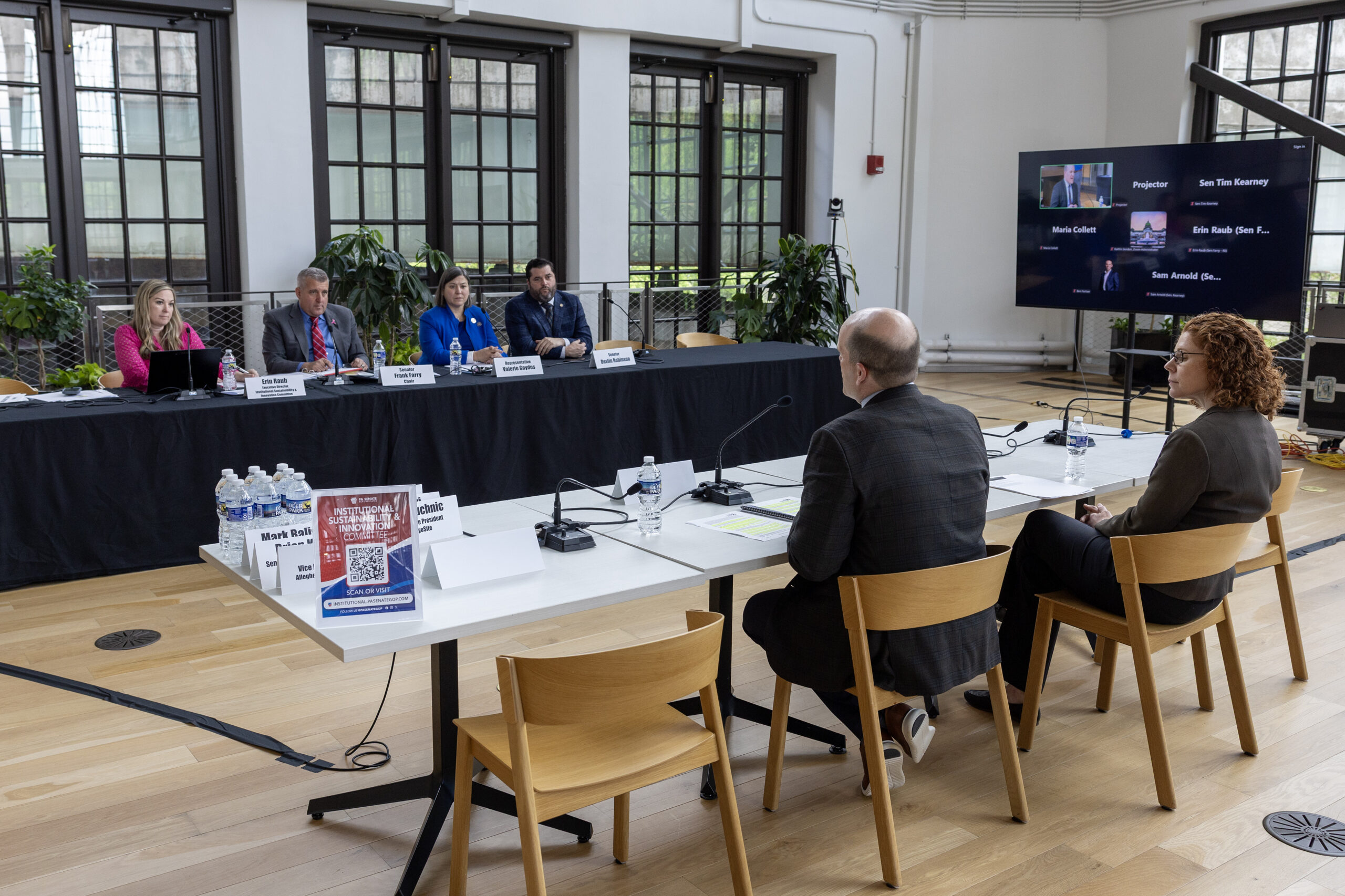 Panel discussion in a conference room with five people seated at a long table facing two speakers, with a screen showing virtual participants in the background.