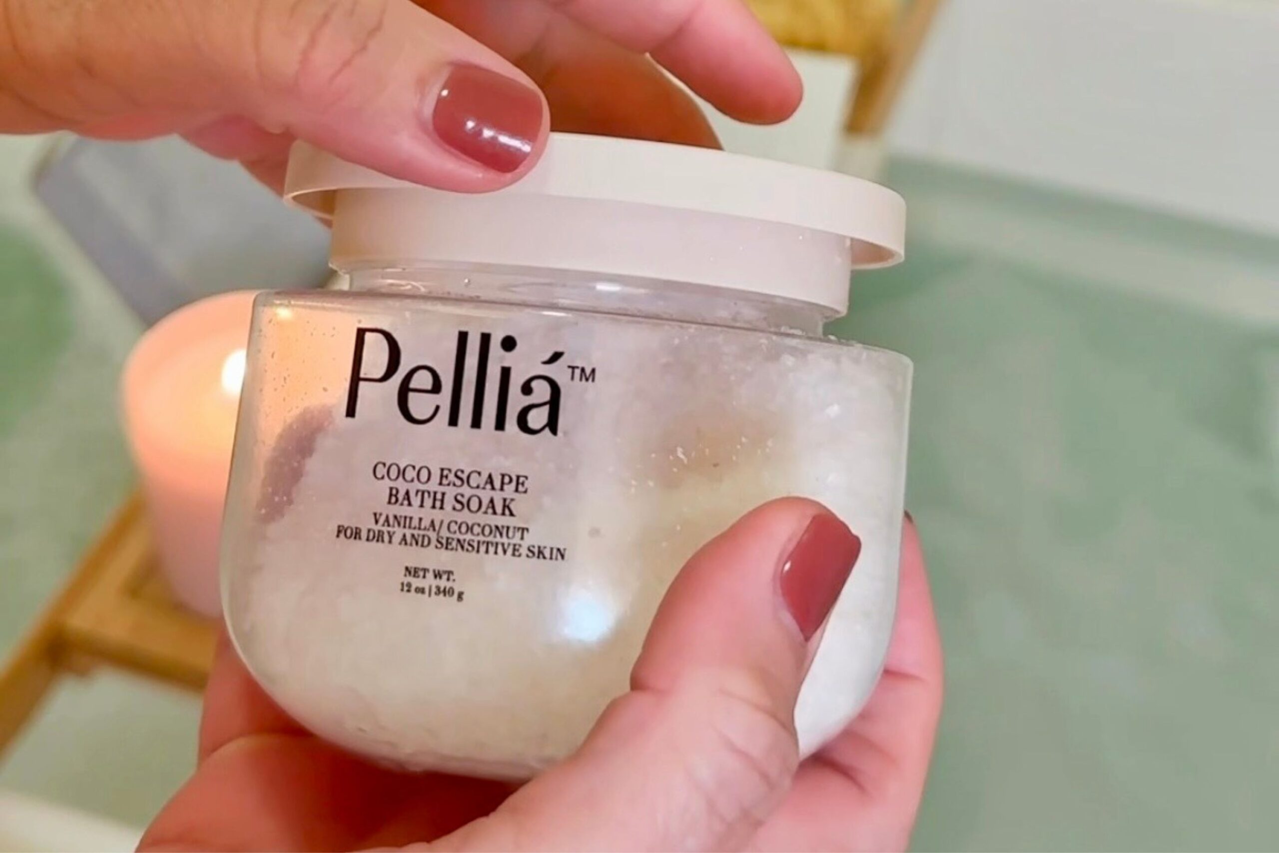 A person with painted nails holds a jar labeled “Pellia Coco Escape Bath Soak for dry and sensitive skin.” A lit candle is blurred in the background.