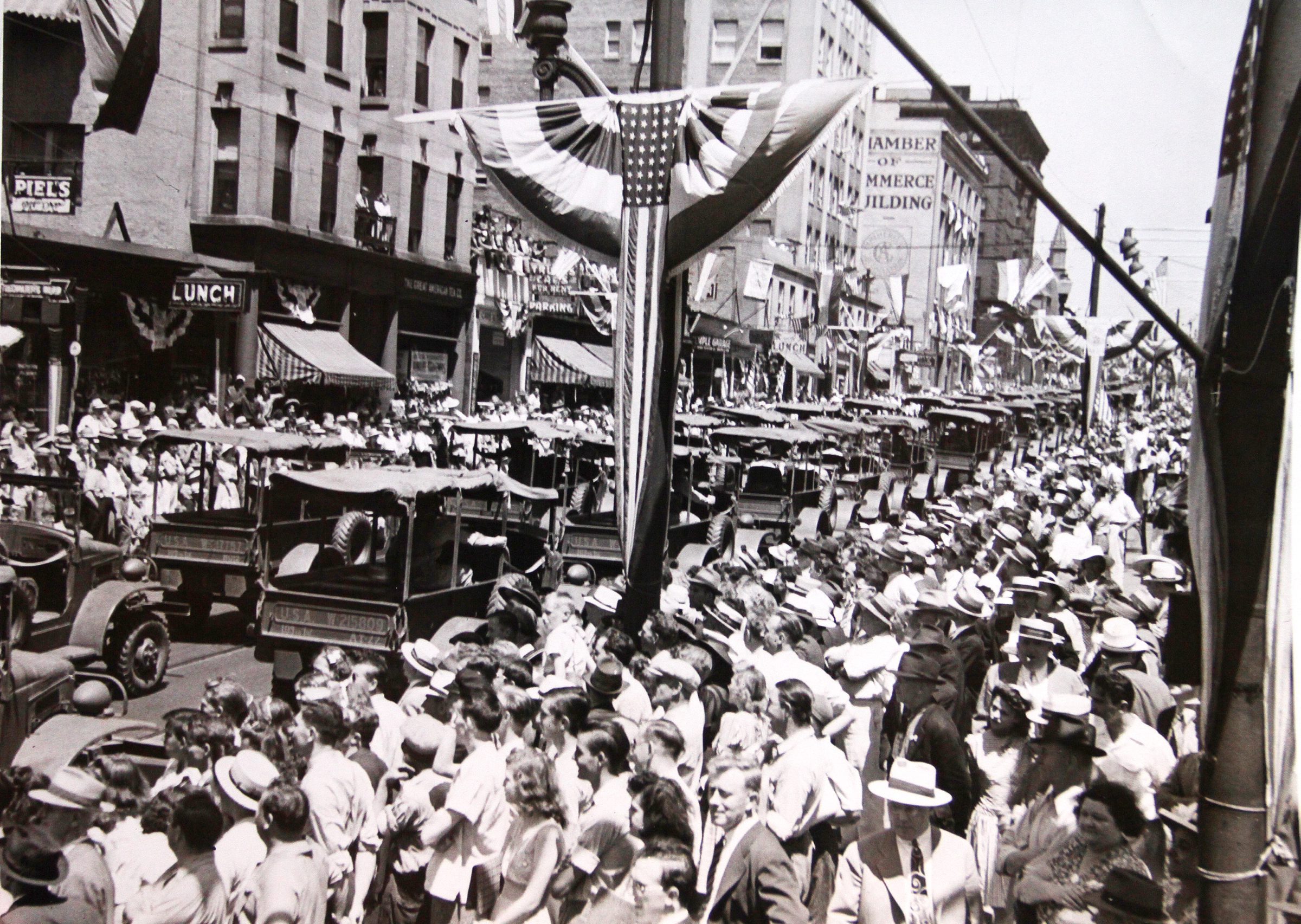 A crowd gathers to watch a procession of Jeeps travel...