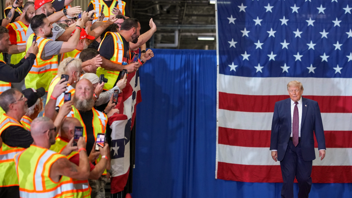 President Donald Trump walks onto a stage in front of a large American flag at the Mack Trucks facility in Macungie, Pennsylvania, while workers in safety vests and supporters take photos and cheer from the crowd.
