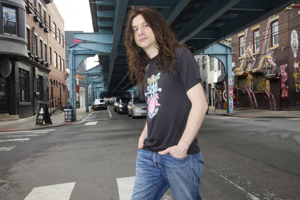 A person with long hair stands confidently on a street under an elevated train track.