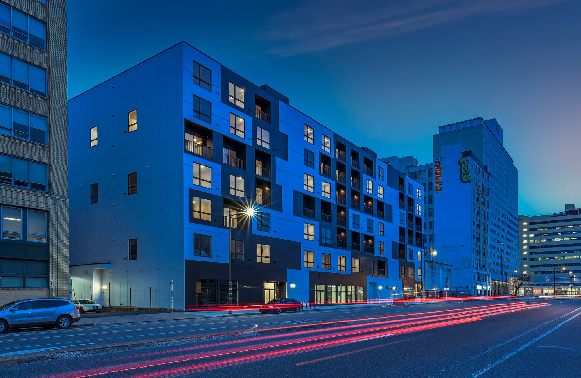 Modern mid-rise apartment building at dusk with illuminated windows, surrounded by city streets and light trails from passing vehicles.