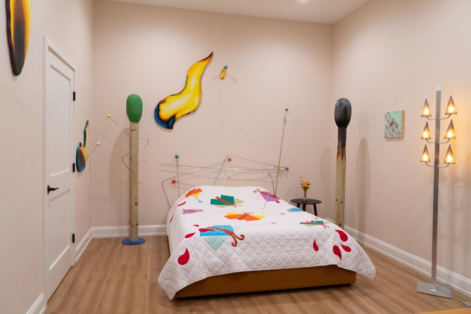 An art-filled bedroom shows a bed with a patterned white quilt, playful wall-mounted sculptures, tall freestanding objects and a lit floor lamp shaped like stacked triangular shades.