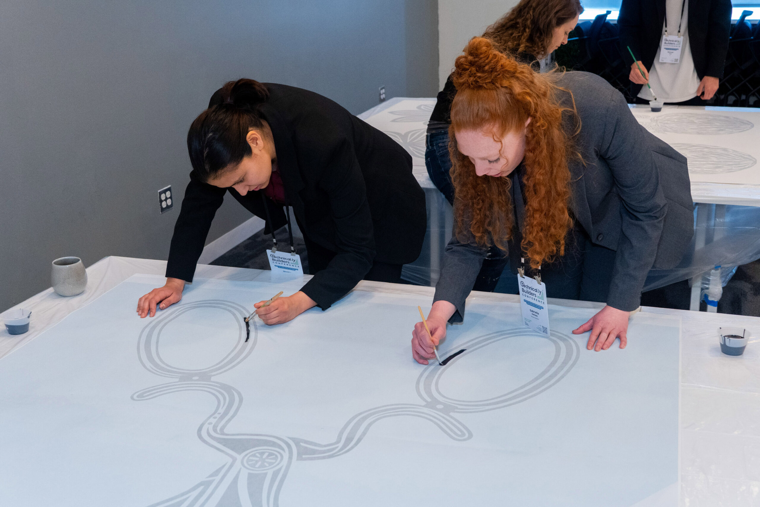 Two women in business attire use brushes to draw intricate patterns on large sheets of paper laid out on a table during an indoor event.
