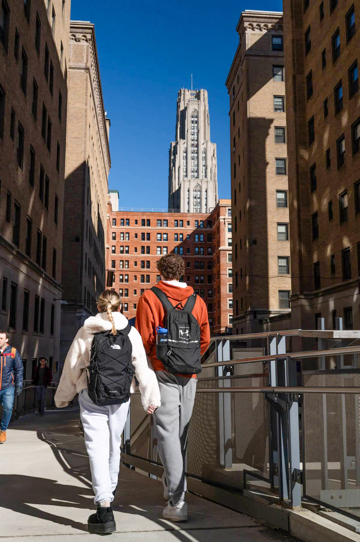 Two people with backpacks walk between tall brick buildings on a sunny day, with a taller skyscraper visible in the background.