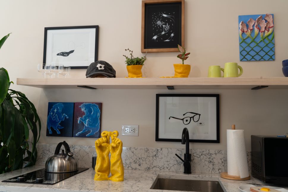 An art-filled kitchenette shows framed works, small paintings, yellow ceramic objects, plants, cups and a yellow hand sculpture arranged around a marble counter and open shelf.