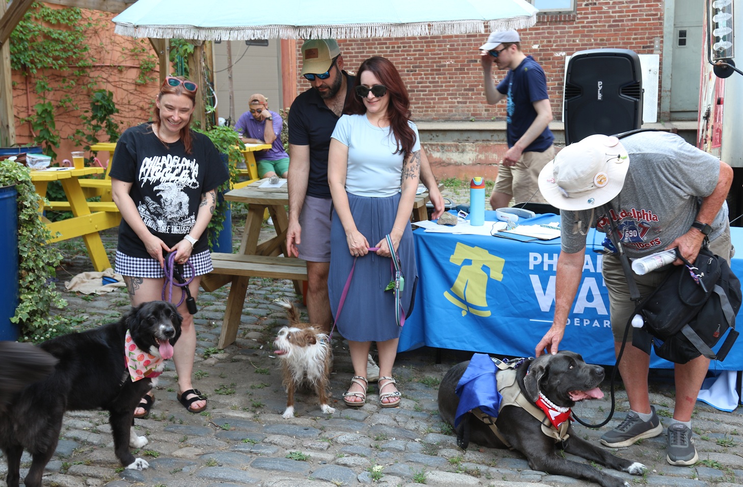 A black dog with a white snout and a happy expression, a brown-and-white dog with slightly frizzy fur, and a large dark gray dog with a content expression hang out with their owners on a cobblestone patio in front of a table draped in a Philadelphia Water Department tablecloth