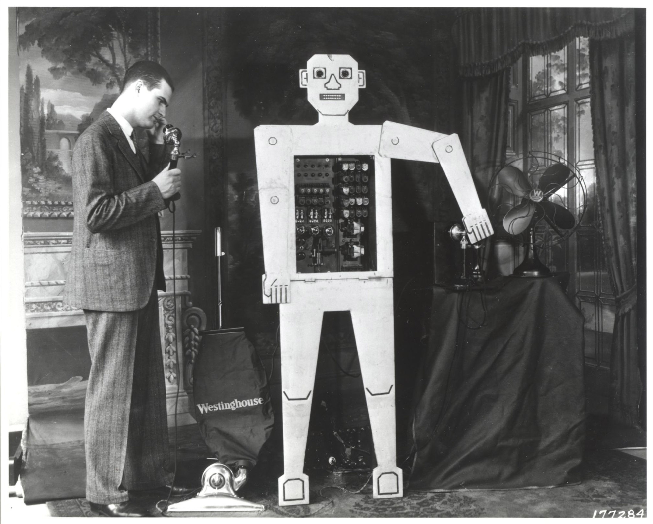 A man speaks into a phone next to an early humanoid robot with visible internal components, standing in a room with a Westinghouse bag and an electric fan.