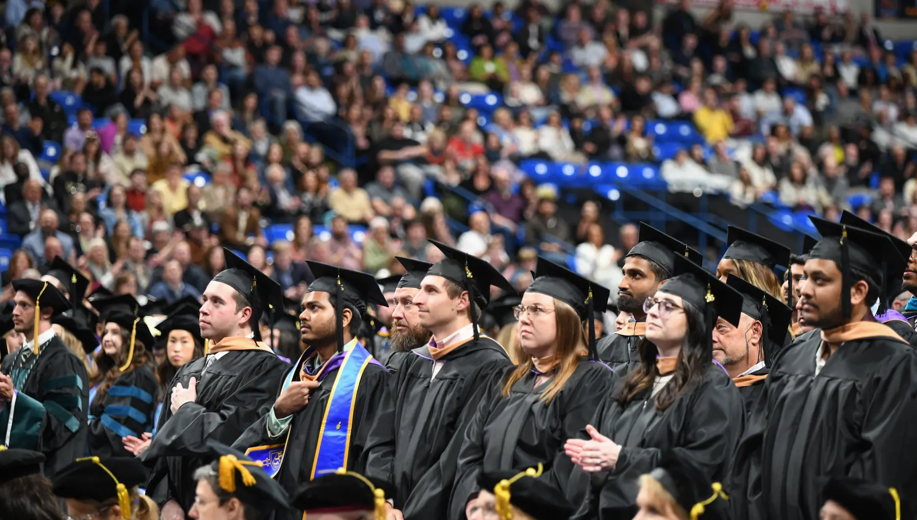 Graduates in caps and gowns stand in rows during a commencement ceremony, with a large audience seated in the background.