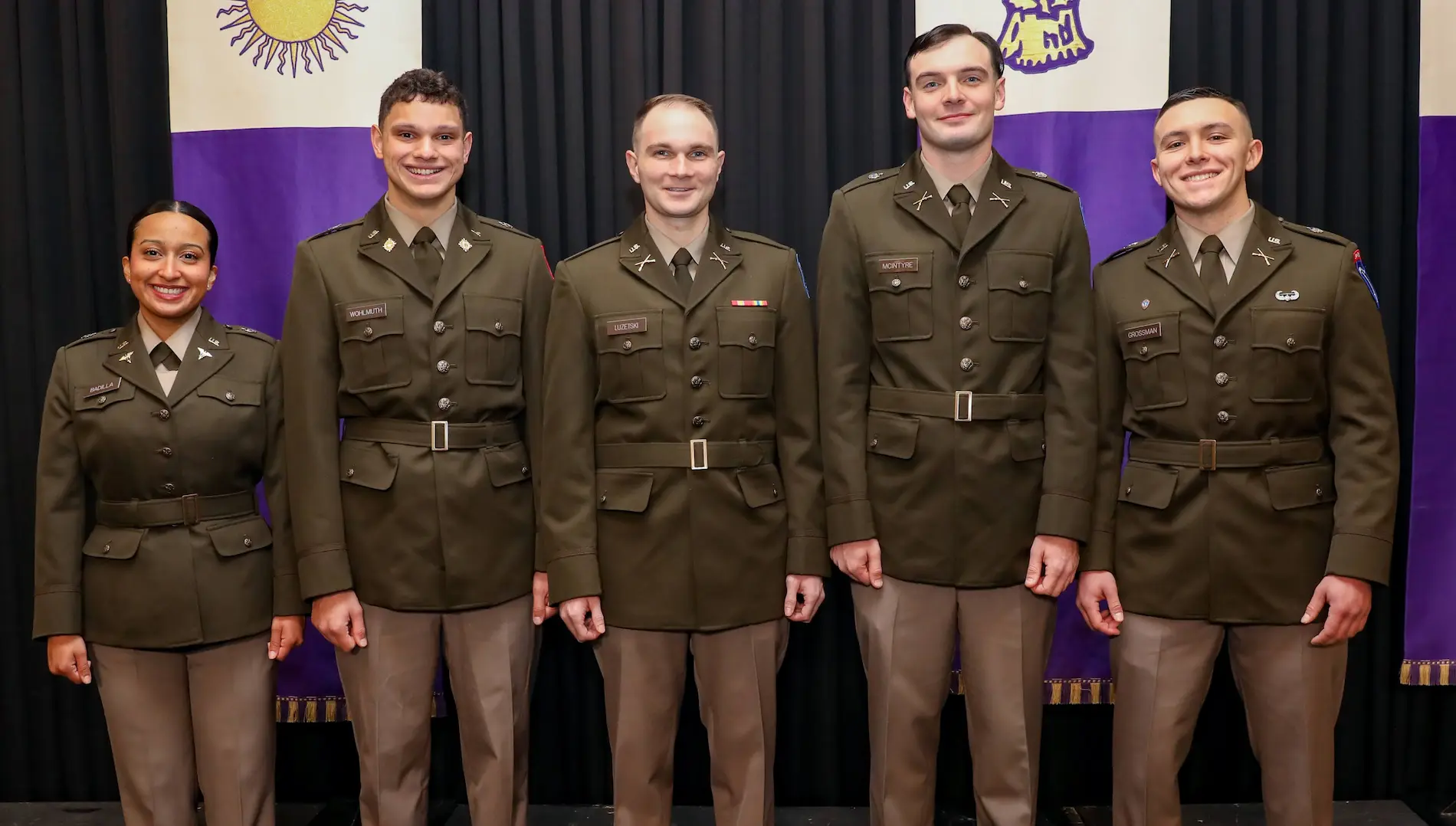 Five U.S. Army cadets in dress uniforms pose and smile in front of purple-and-white banners.