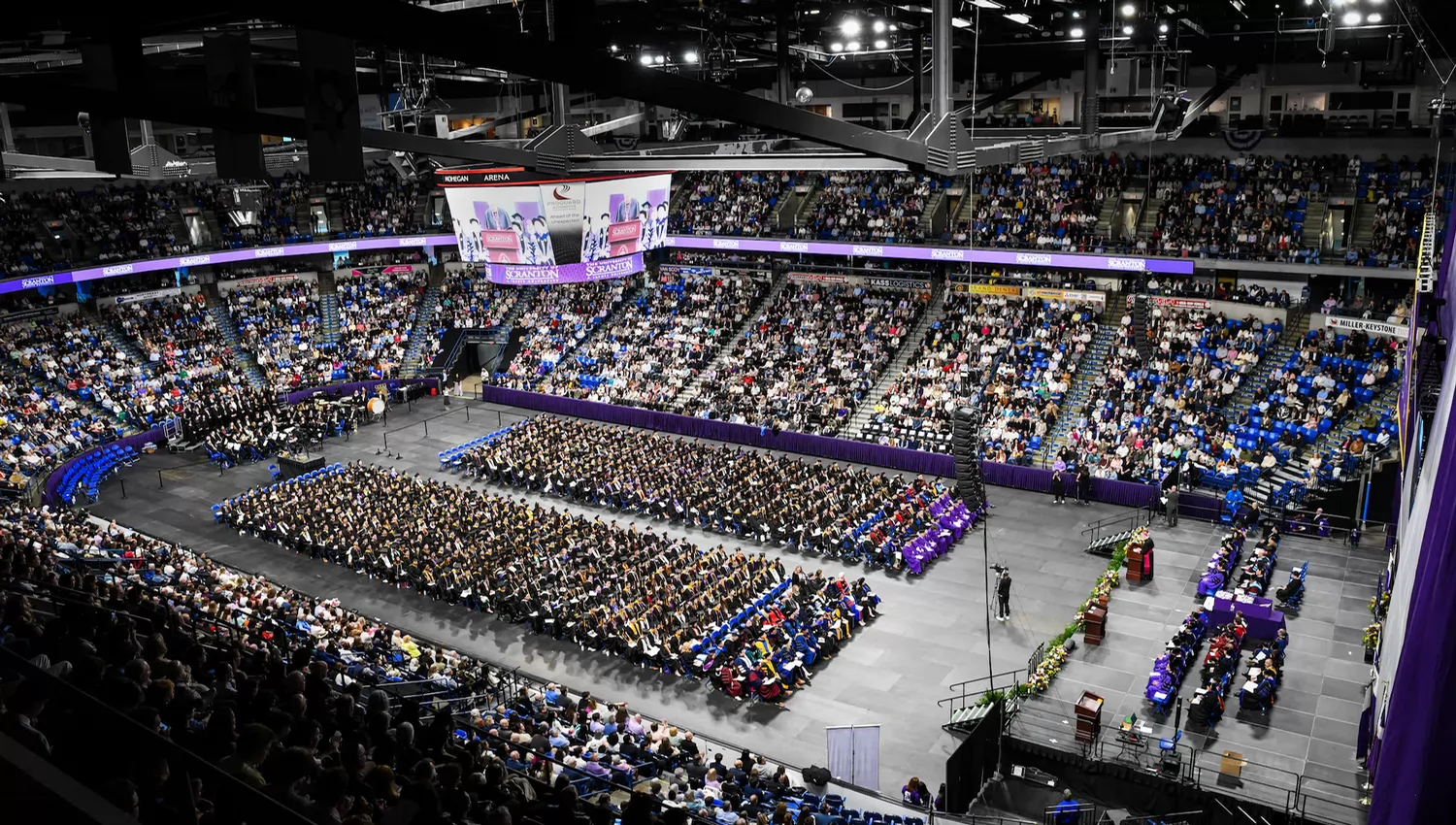 Wide view of a graduation ceremony in a packed indoor arena, with rows of graduates seated on the floor and a stage set up at the front.