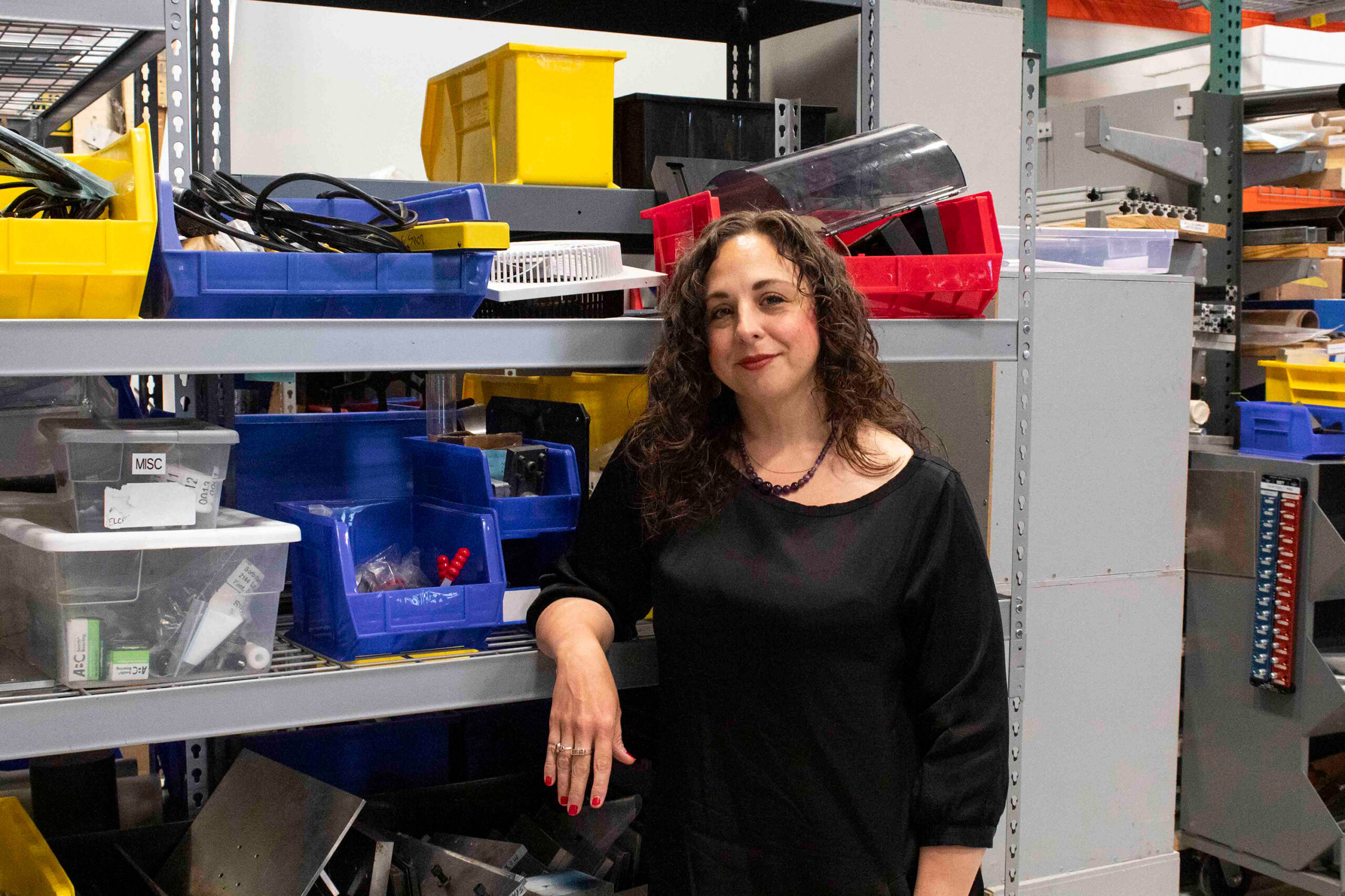 A woman in a black shirt stands beside metal shelves filled with plastic bins and various hardware items in an industrial storage area.