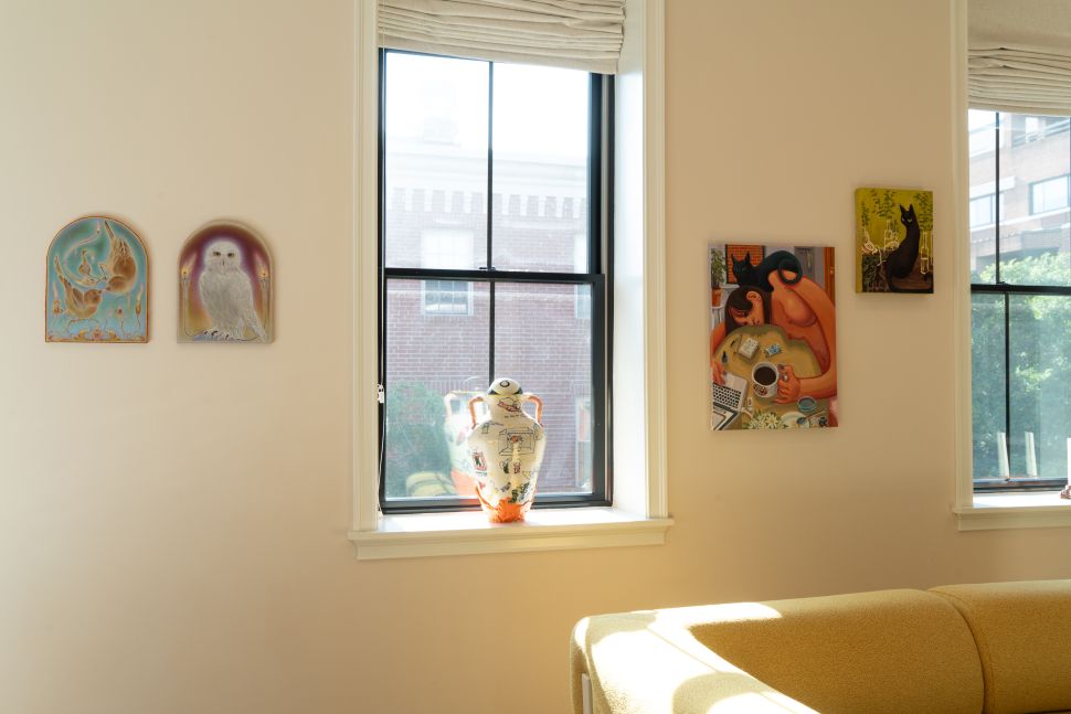 An interior wall near two windows displays small paintings, arched artworks and a ceramic vase on the windowsill above the back of a yellow sofa.