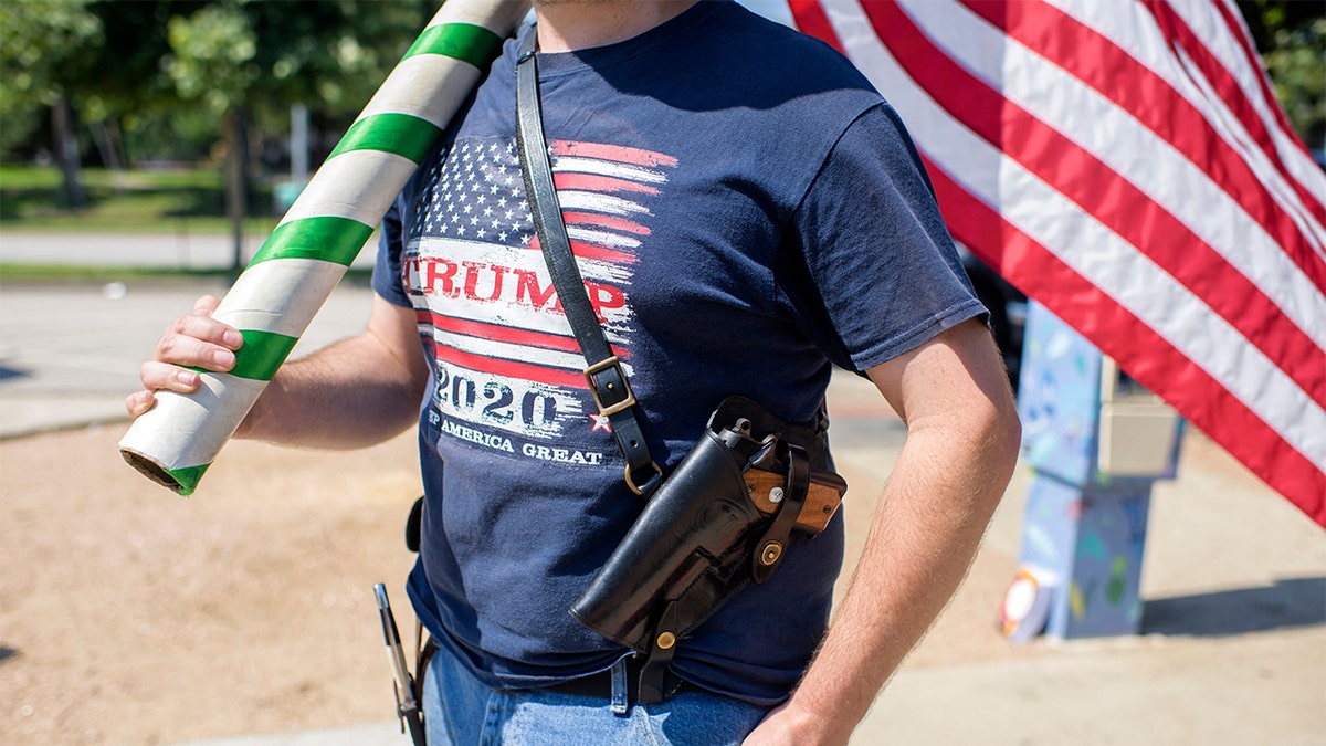 A person wearing a Trump 2020 T-shirt carries a holstered handgun and an American flag while standing outdoors at a pro-gun rights event.