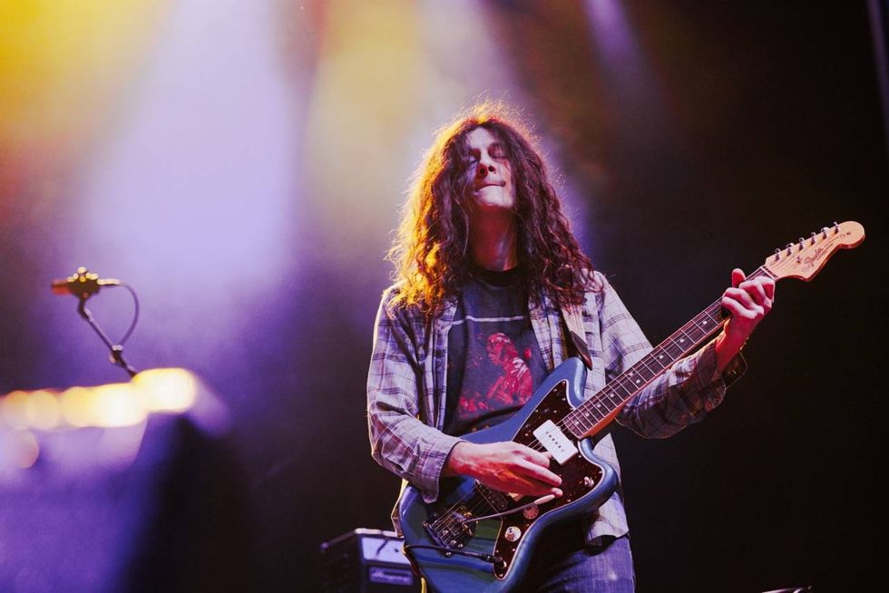 Musician with long hair plays a blue guitar on stage, bathed in colorful lights.