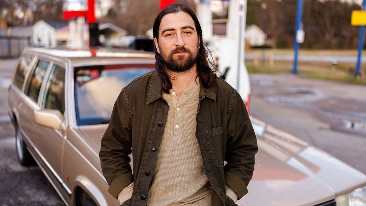 Noah Kahan poses by brown wagon