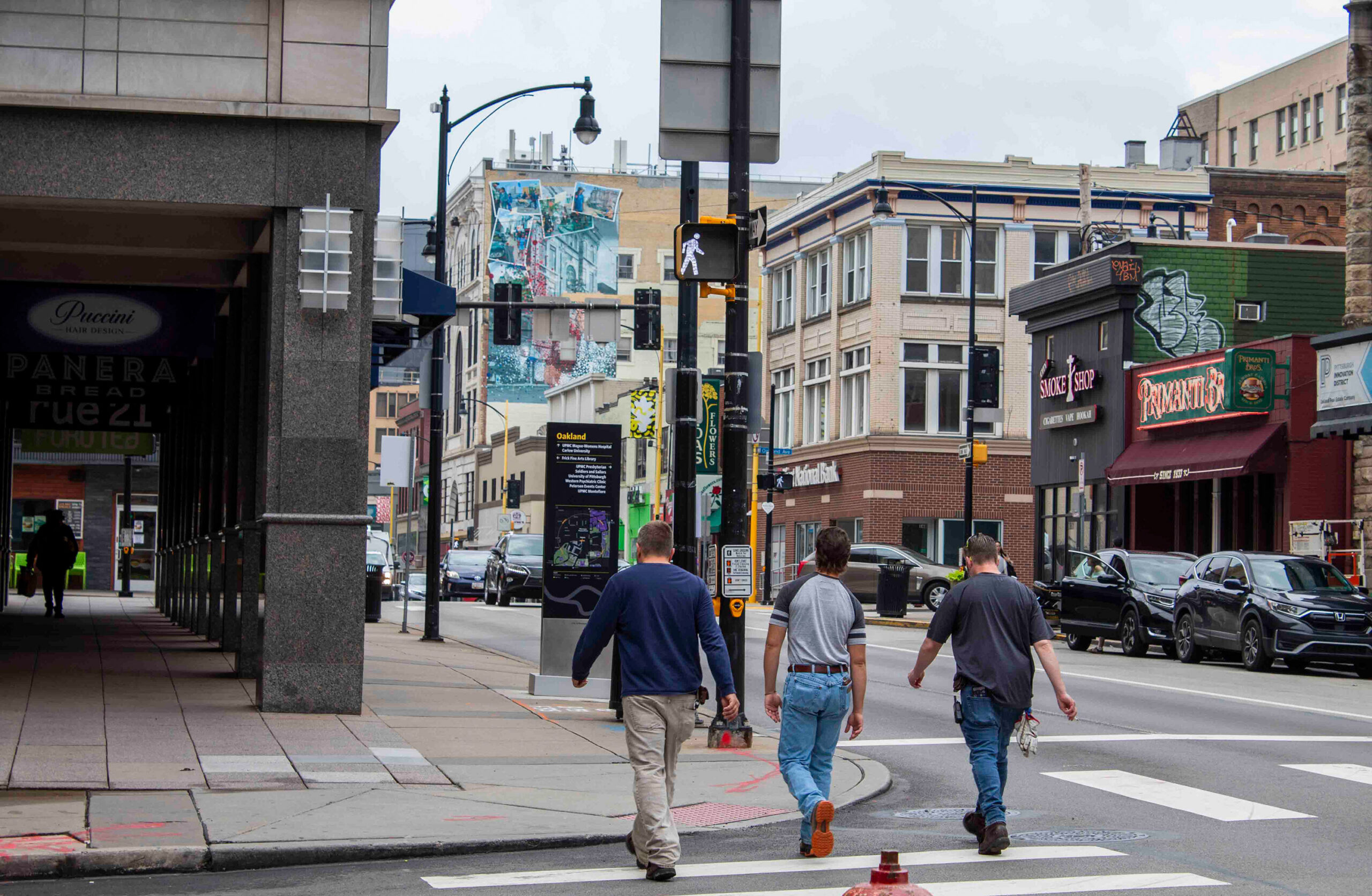 Three men walk across a city street at a crosswalk lined with restaurants and shops on an overcast day.