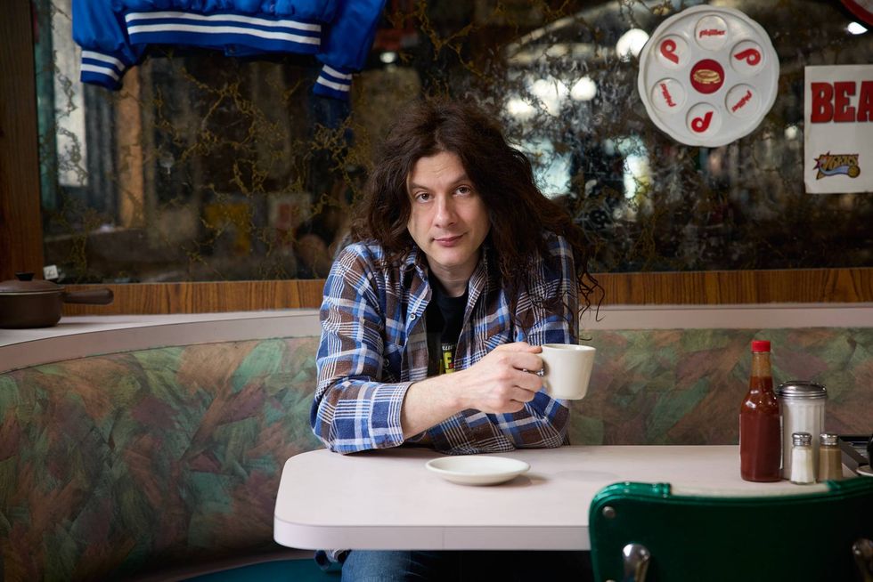 Person with long hair sits at a diner table, holding a coffee cup, casual plaid shirt.