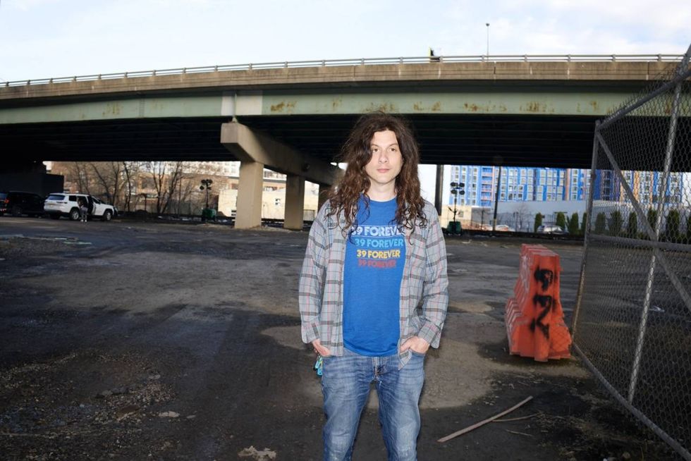 Person with long hair stands on a parking lot under an overpass, wearing a blue shirt and plaid jacket.