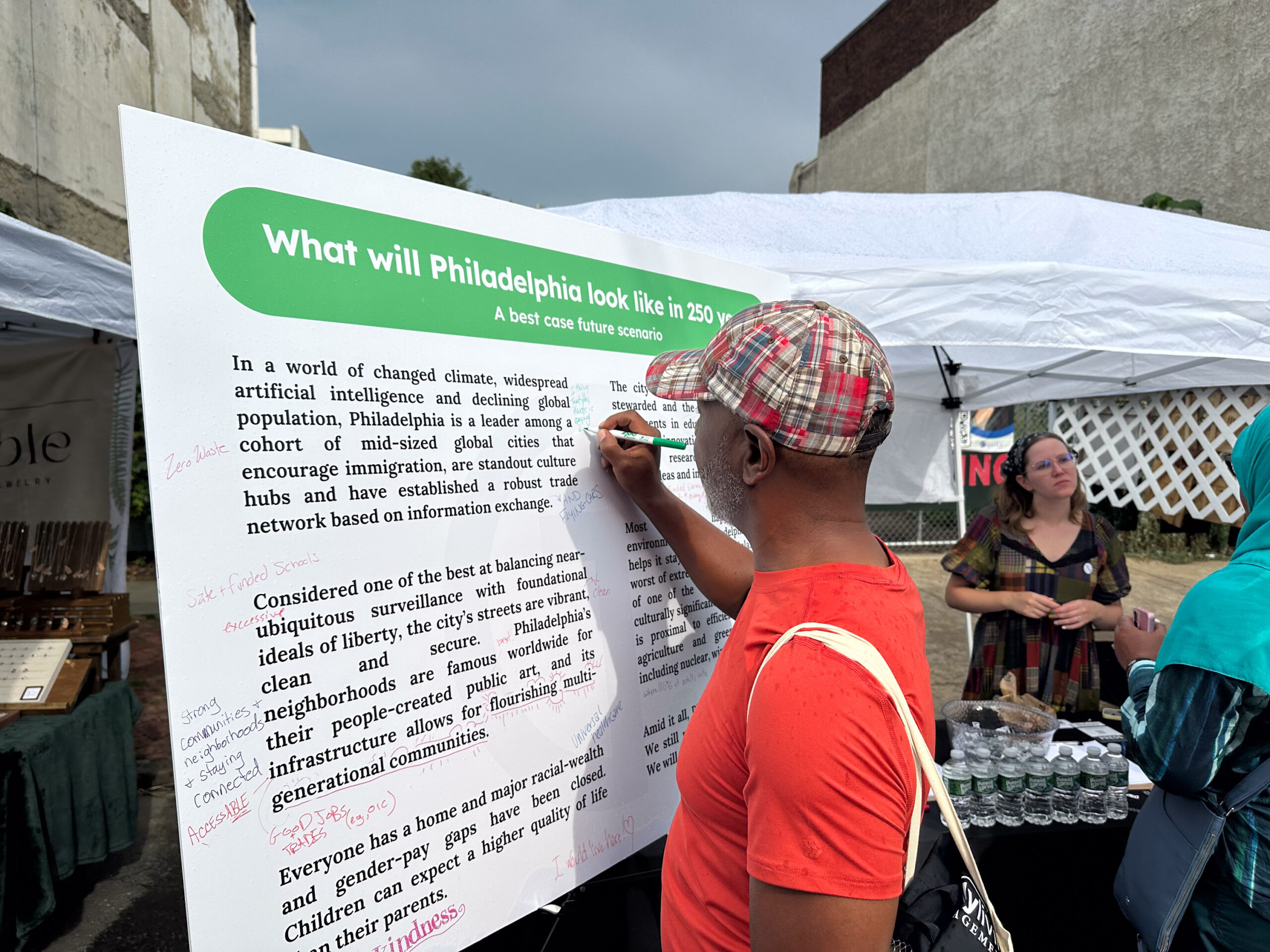 A man writes on a large display board about Philadelphia's future at an outdoor event, while two women stand nearby under a white tent.