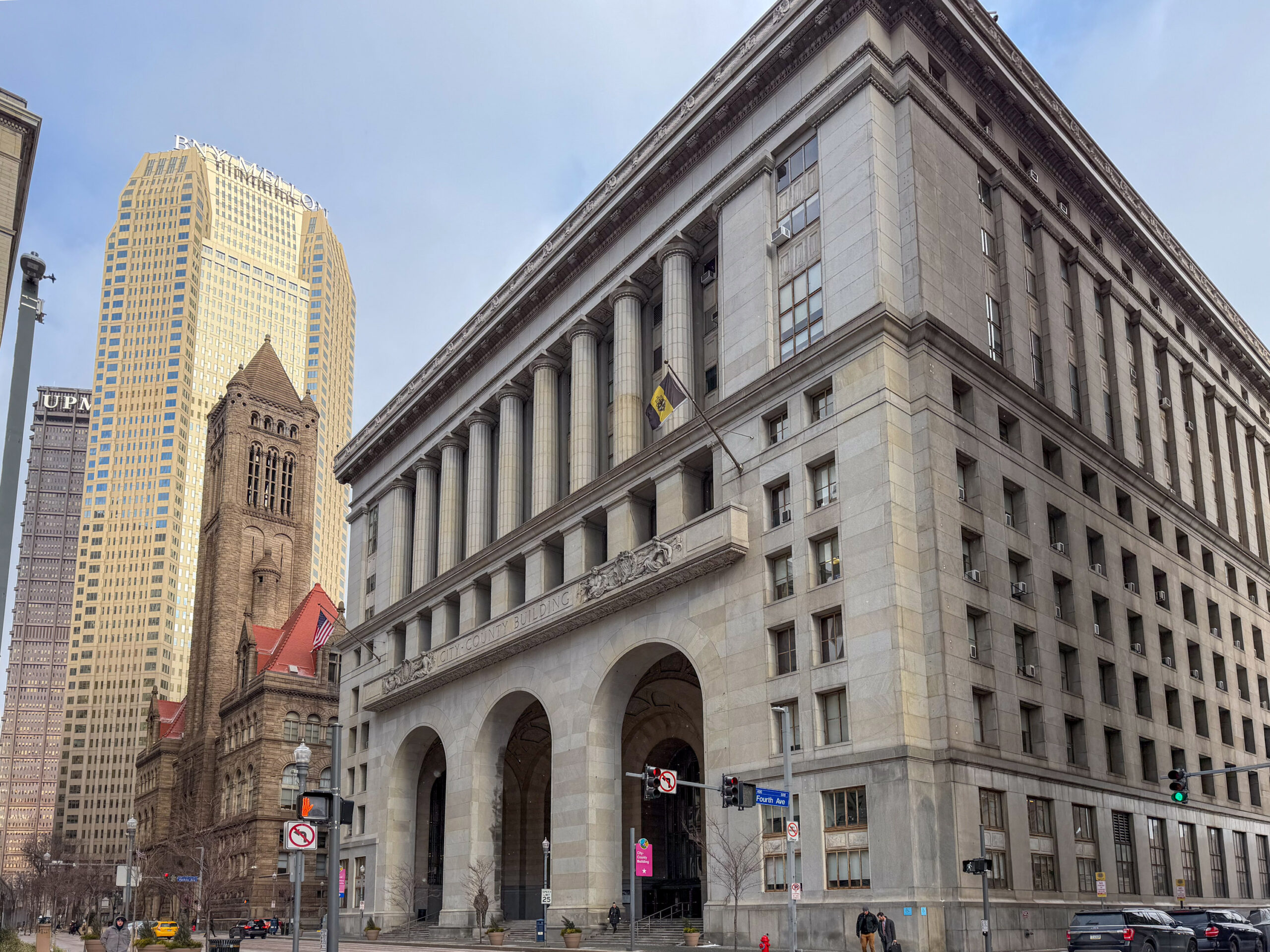A large stone government building with columns and arches stands on a city street corner, with modern and historic buildings visible in the background.