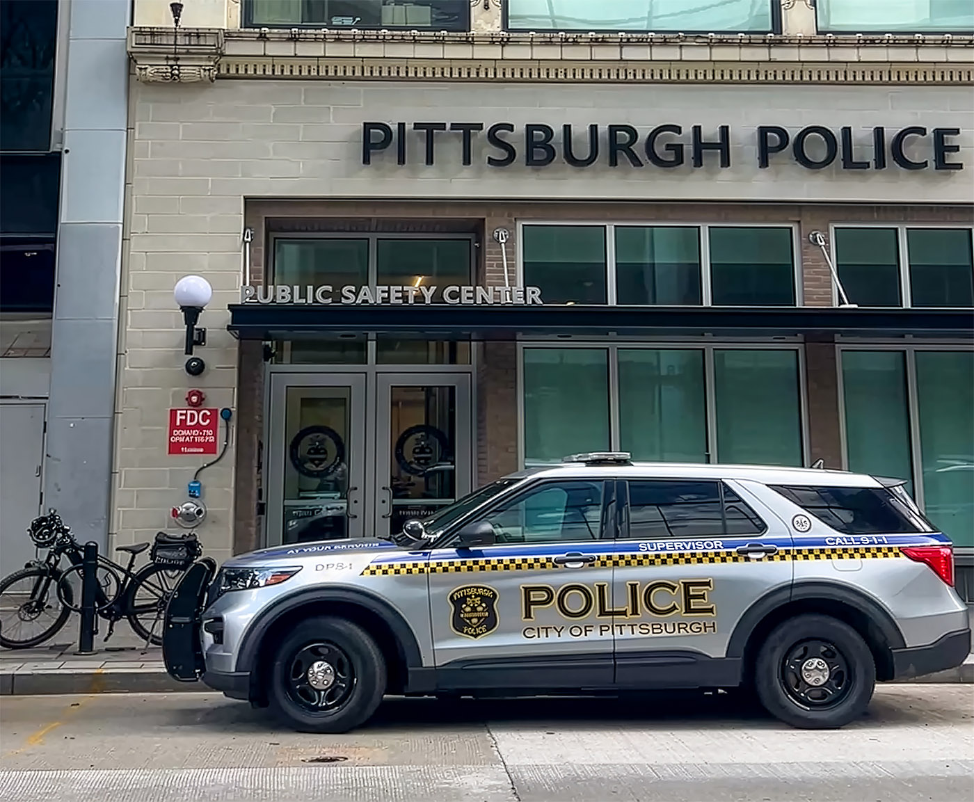 A Pittsburgh Police SUV is parked outside the Public Safety Center building in downtown Pittsburgh. Two bicycles are locked to a bike rack nearby.