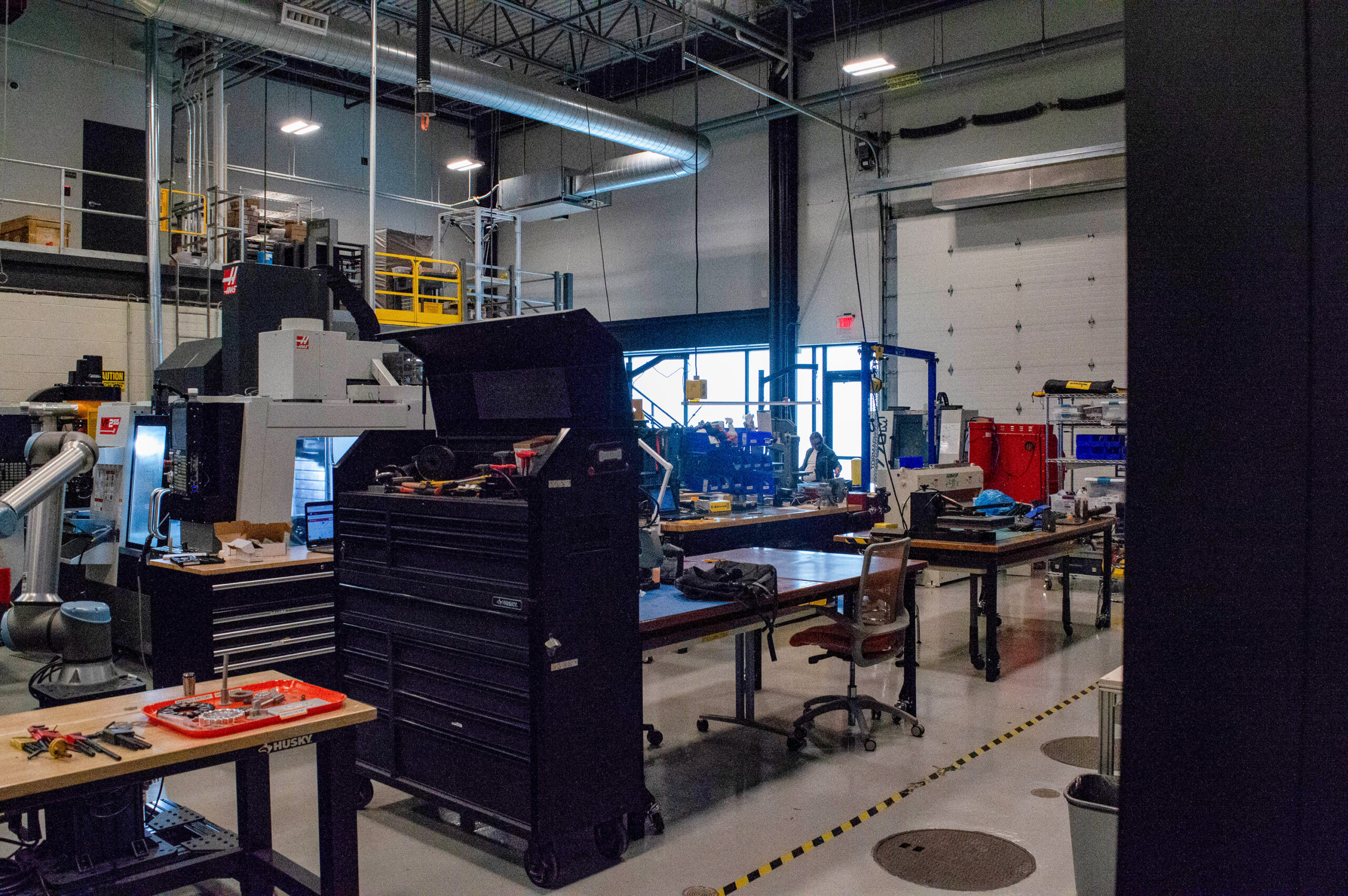 A workshop with toolboxes, workbenches, equipment, and various tools. The area is well-lit with industrial lighting and has a garage-style door in the background.