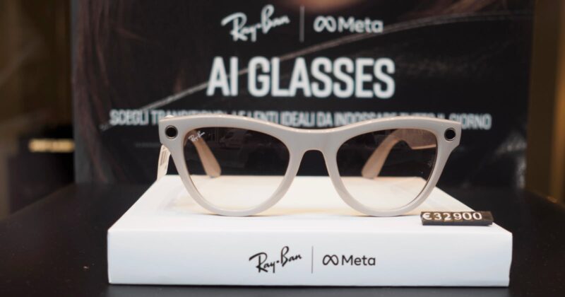 A pair of Ray-Ban and Meta AI glasses is displayed on a white stand with the price tag €329.00. The background features branding for Ray-Ban and Meta with the text "AI GLASSES.