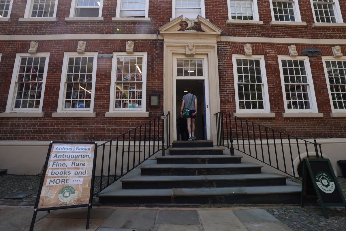 A man walks in through the front door of Aldous Books in Bluecoat Chambers in Liverpool City Centre. He is wearing white trainers, dark blue shorts and a grey shirt. He has a large white bag slung across his body