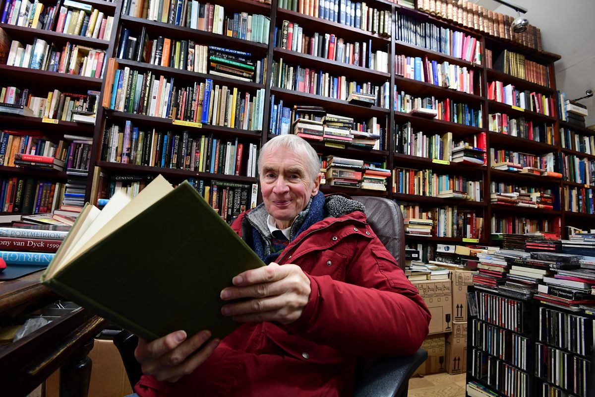 A man sits in a chair inside a bookshop. Surrounded by shelves stacked high with books, he's holding a back himself. He's wearing a red coat and is smiling toward the camera. He has grey, thinning hair.
