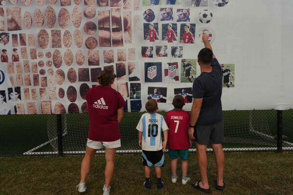 Pennsylvania Classic coaches, Brittney Jakobson, left, and Nick Jakobson, right, look at a banner of U.S. national team soccer player Christian Pulisic with their children, Declan Jakobson, who wears an Argentina jersey, and Camden Jakobson, wearing a Portugal jersey, at the club were Pulisic honed his skills in Manheim, Pa., on Monday, June 29, 2026. (AP Photo/Luis Andres Henao)