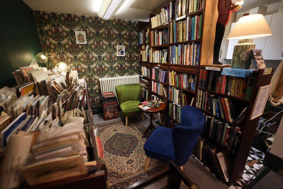 A cosy looking room lined with shelves and free standing bookshelves. They are filled with books of all descriptions. There is a small occasion table on the right hand side of the room, with two plush covered chairs sat either side of it. One chair is a rich, navy blue, the other an olive green. Behind the green chair stands a radiator. The back wall of the room is papered with a richly patterned wallpaper in greens, reds and browns. There a free standing lamps dotted around the room which provide ambient lighting