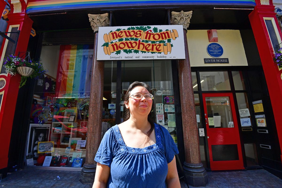Maria Ng stands outside News From Nowhere in the sunshine on Bold Street. She's wearing glasses and has her dark hair pulled back off her face. She's wearing a blue top with elbow length sleeves