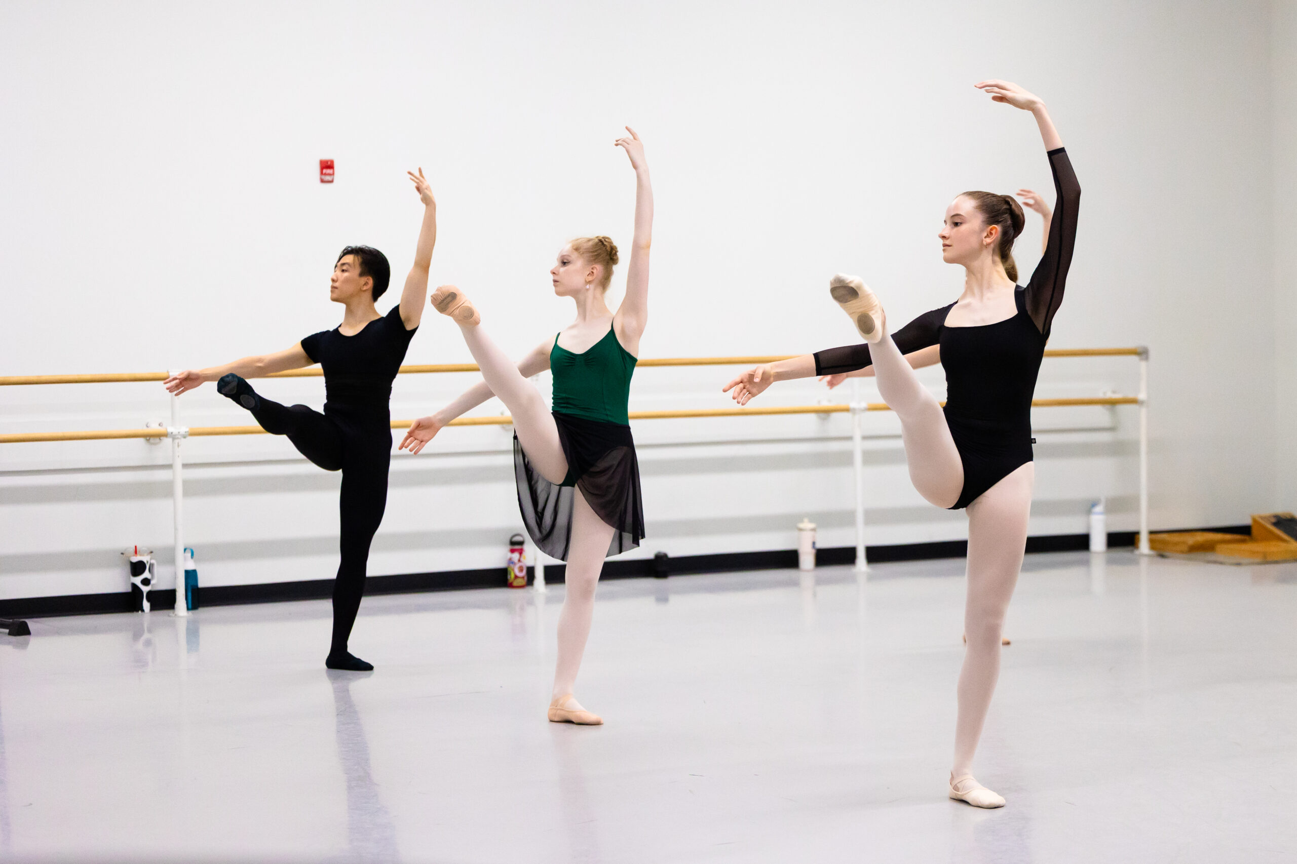 A male and two female ballet students pose in développé croisé devant during a ballet class in a large dance studio.