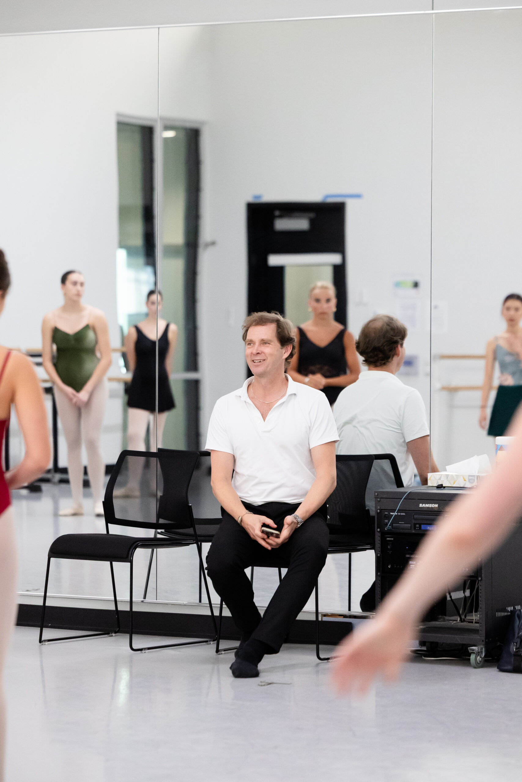 A male ballet teacher wearing a white polo shirt and black pants sits in a chair at the front of the dance studio and smiles as he looks out at his ballet students.