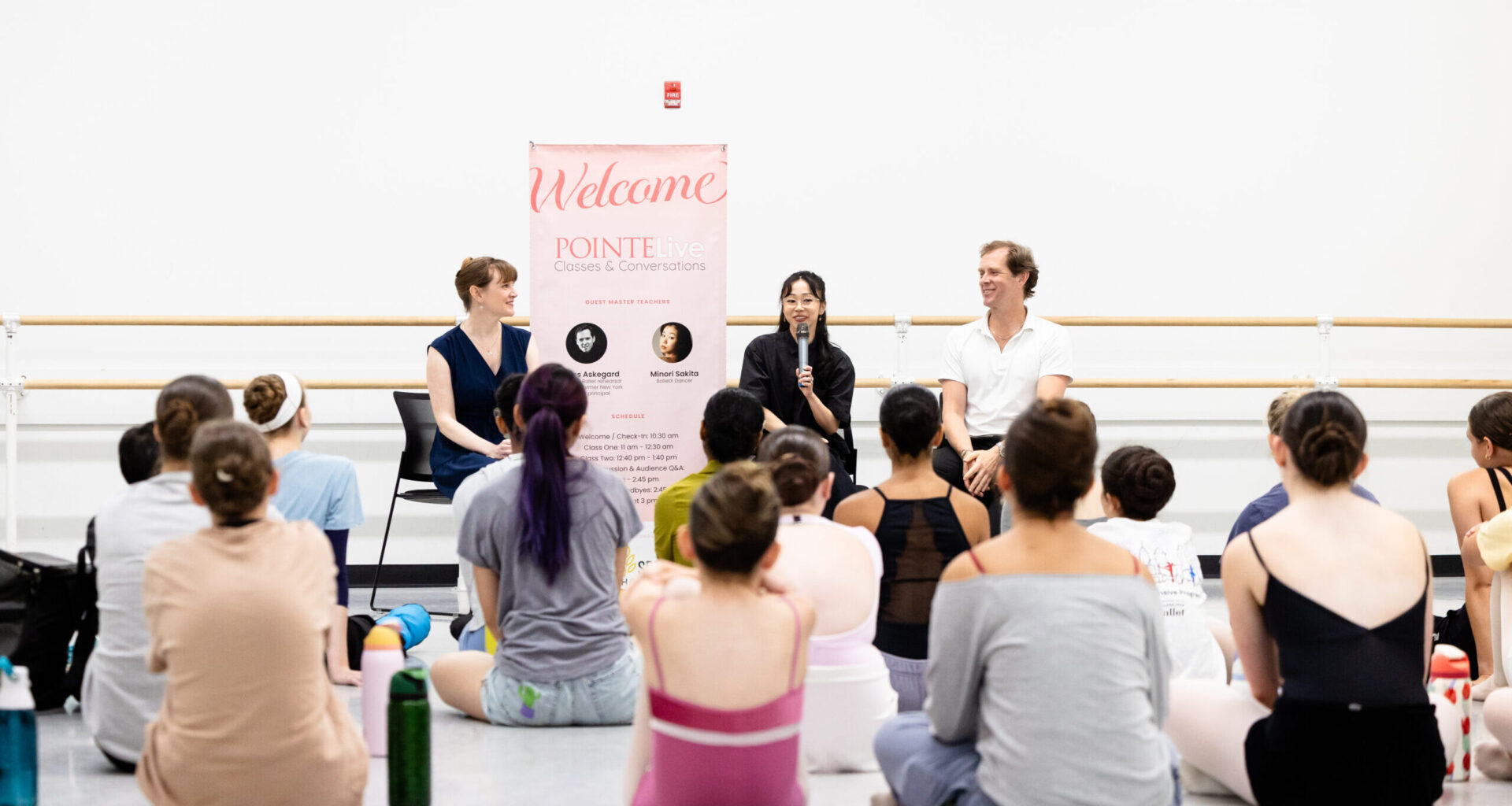 Two women and a man sit in black chairs against the wall of a large dance studio in front of a group of dancer students who wear rehearsal clothing. The woman in the middle holds a microphone and addresses the dancers.