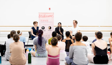 Two women and a man sit in black chairs against the wall of a large dance studio in front of a group of dancer students who wear rehearsal clothing. The woman in the middle holds a microphone and addresses the dancers.