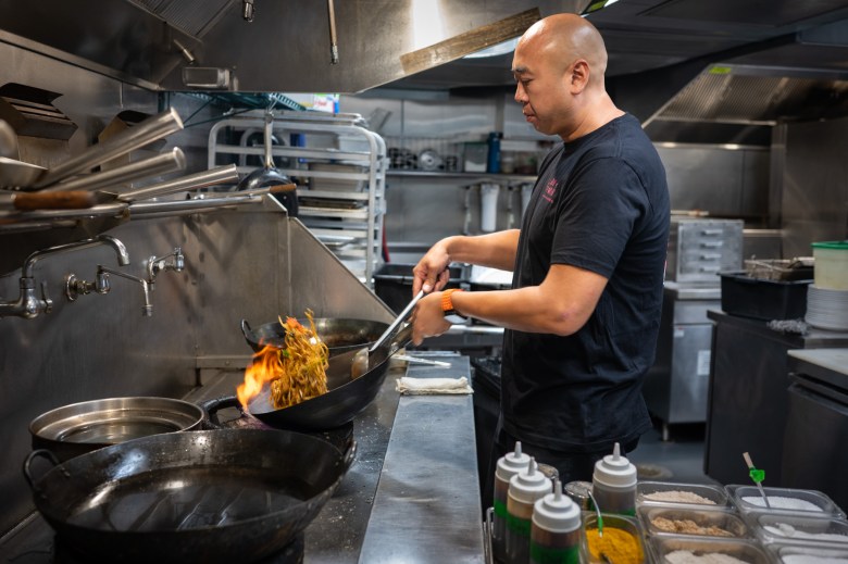A chef in a commercial kitchen cooks noodles in a wok over an open flame, surrounded by cooking utensils and ingredients.