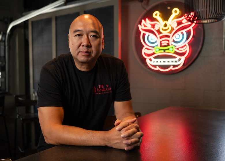 A man with a shaved head sits at a counter, arms folded, wearing a black T-shirt. A colorful neon dragon sign is illuminated on the wall behind him.