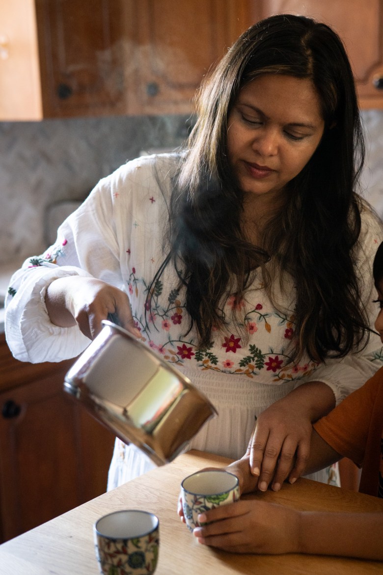 A woman in a white floral dress pours a hot beverage from a metal pot into a cup while a child sits beside her at a wooden table.