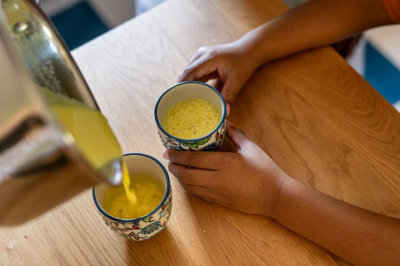 A person pours a yellow beverage from a pot into a patterned cup on a wooden table, holding another filled cup in their other hand.
