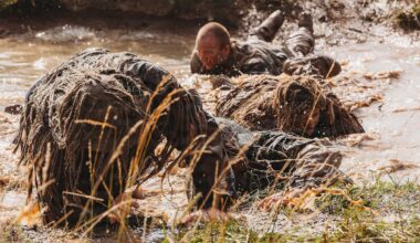 Four people in camouflage military uniforms crawl through a muddy stream on their stomachs.