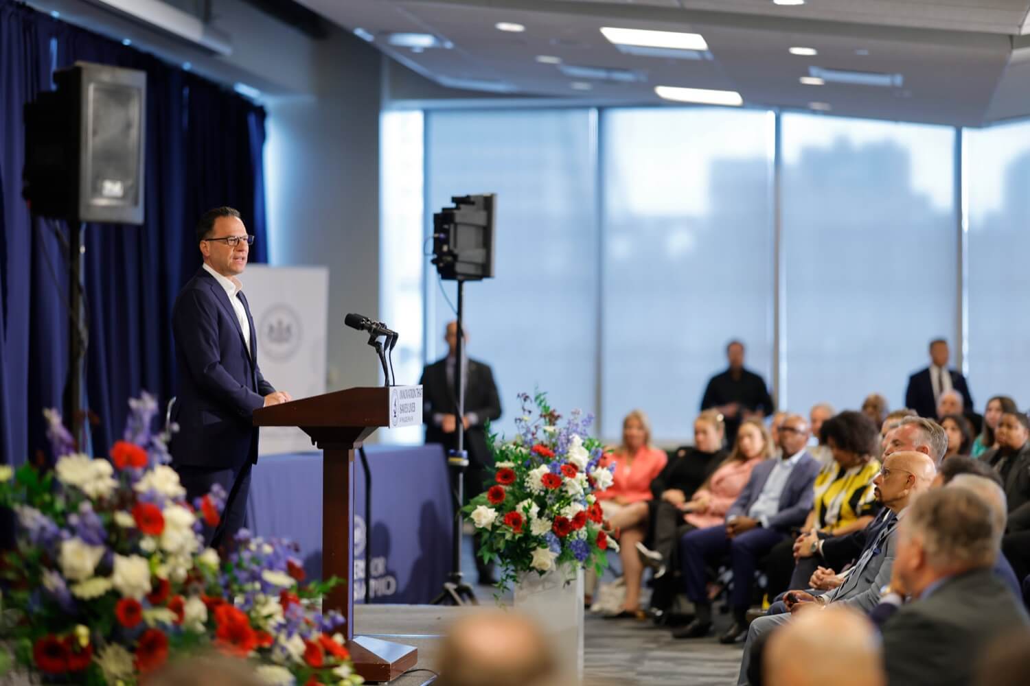 Governor Josh Shapiro in a suit speaking at podium during today's B+labs tour event with seated audience and floral arrangements.