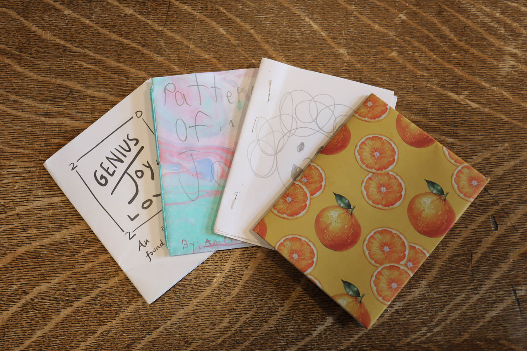 Four handmade booklets with illustrated covers, including one with oranges, are arranged in a fan shape on a textured brown surface.