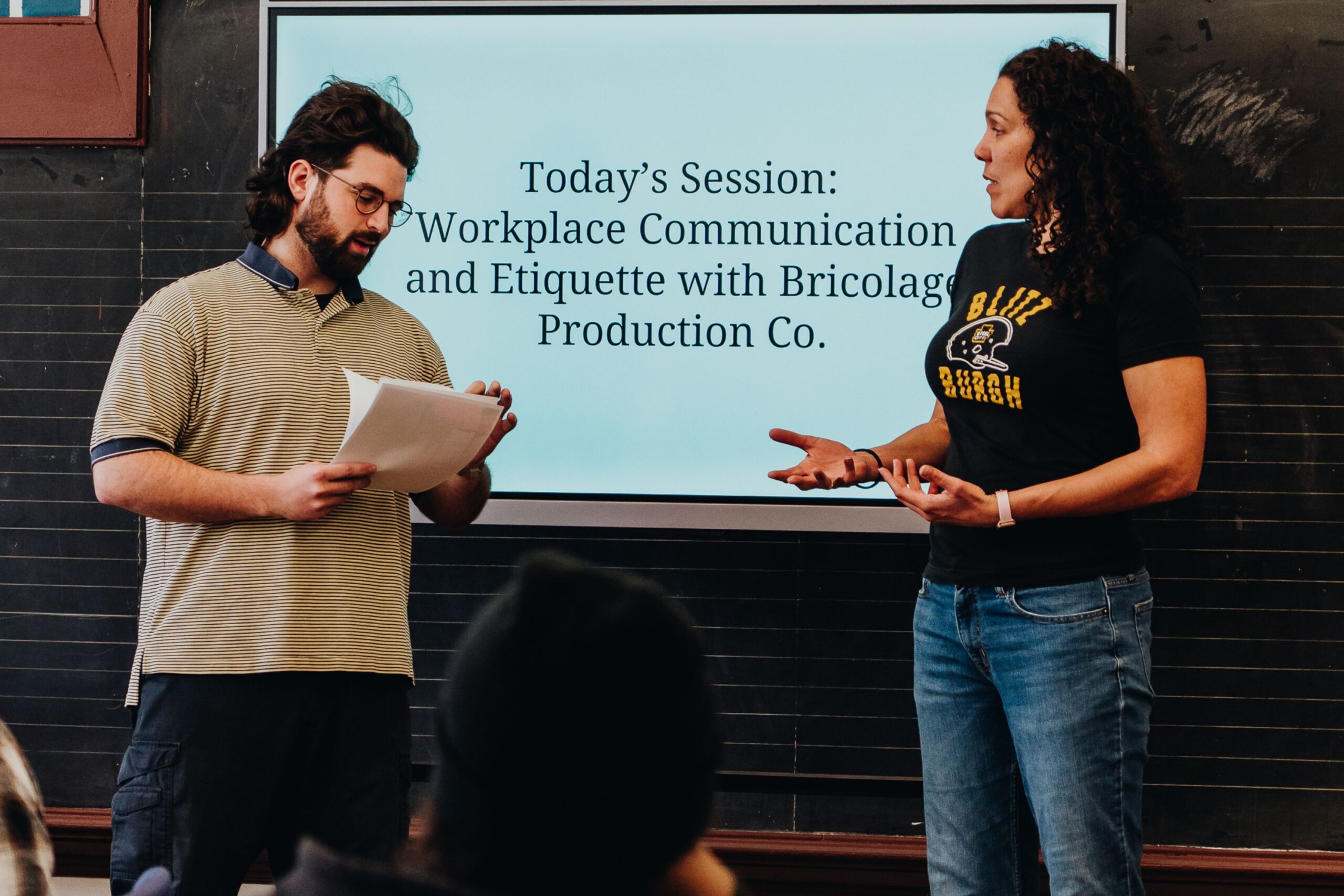 Two people stand in front of a screen that reads "Today's Session: Workplace Communication and Etiquette with Bricolage Production Co." while talking and holding papers.