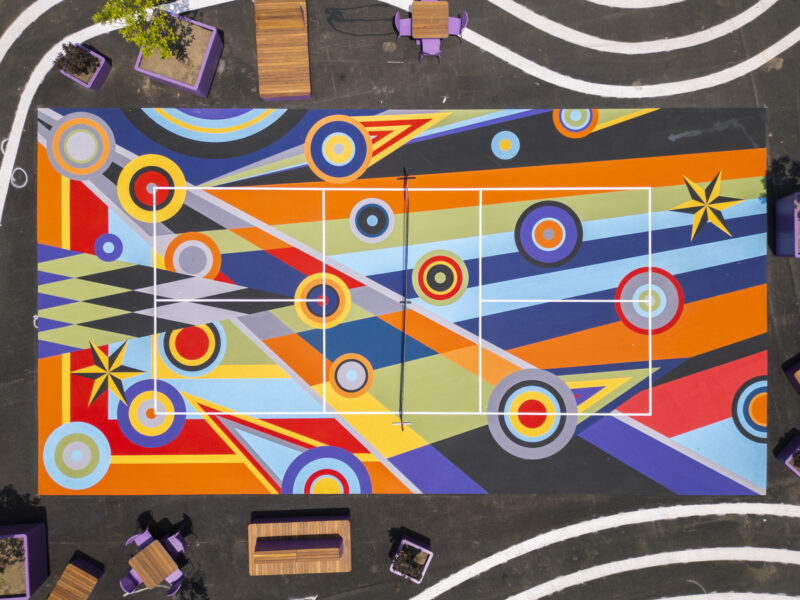 Aerial view of a vividly painted sports court featuring Sharmistha Ray Geometry of Play-inspired geometric patterns in bright colors, surrounded by benches and plants.