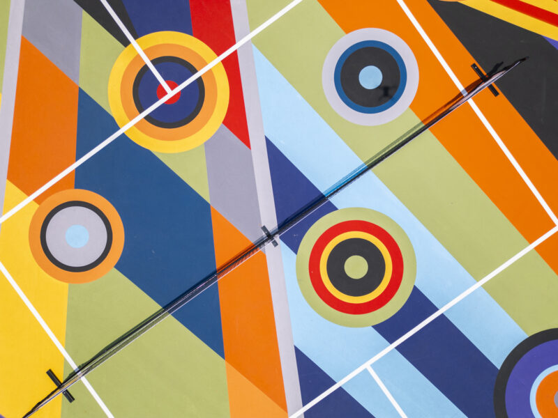 Aerial view of a colorful abstract mural inspired by Sharmistha Ray’s Geometry of Play, with vibrant geometric shapes and concentric circles, intersected by a black pipe and white grid lines.
