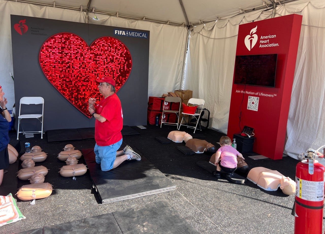 An instructor teaches Hands-Only CPR Training at the FIFA Fan Festival, kneeling next to several dummies on the ground.