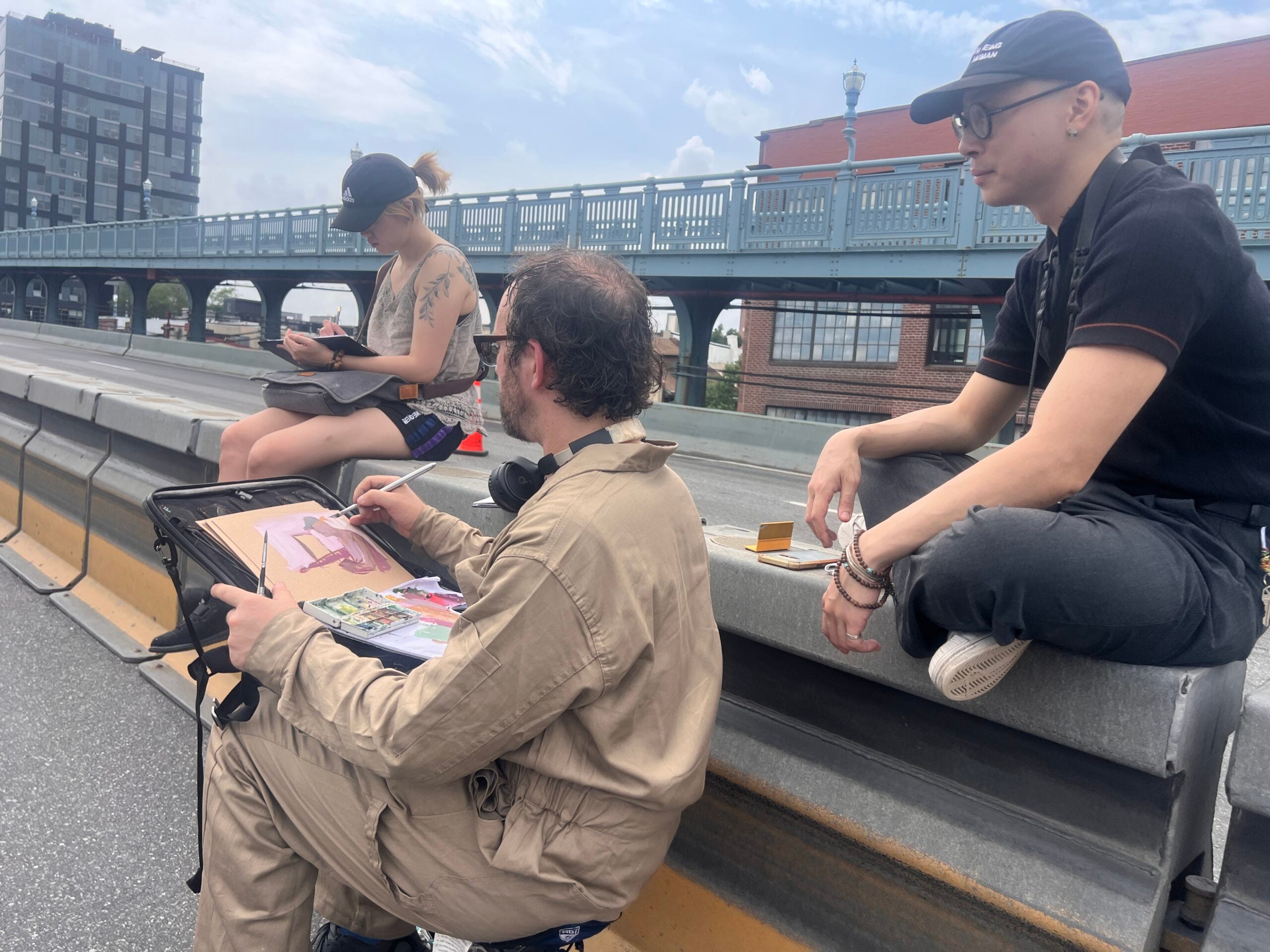 3 people sit together on the Ben Franklin Bridge. One has a paint set and is seemingly painting his other friend on the bridge, who seems to be drawing something too.